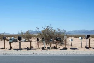 A row of variously styled mailboxes lining a desert road, with sparse vegetation and mountains in the background, under a clear blue sky.